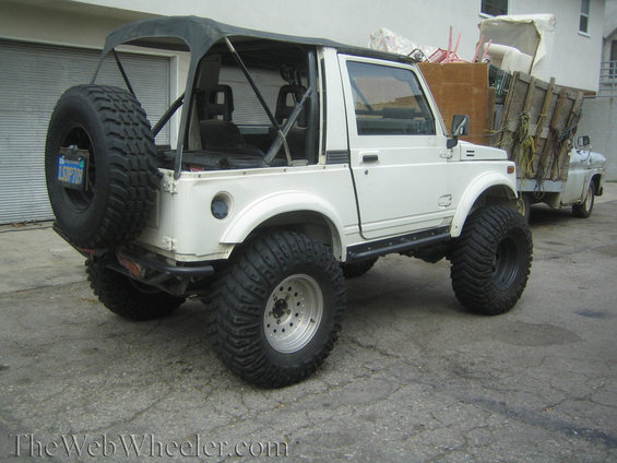 White 1987 Suzuki Samurai with oversized tires and soft top, parked next to a utility truck