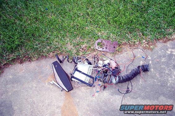 Pile of vehicle wiring, a black taillight assembly, and a rusted metal bracket on concrete next to grass