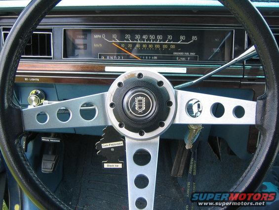 Interior view of a car's dashboard, showing the steering wheel, speedometer, and fuel gauge. A 'Car-Freshener' hangs from the steering column