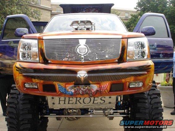 Lifted Cadillac Escalade with custom eagle, flag, and 'HEROES' graphics, and a blower hood scoop. Another vehicle is visible to the left