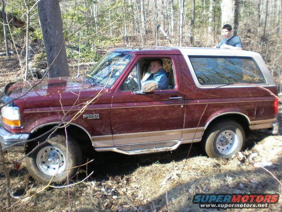Maroon and tan Ford Bronco off-road, driver visible, with a person standing on the roof