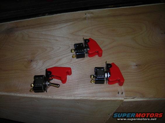 Three toggle switches with red protective covers are laid out on a wooden surface