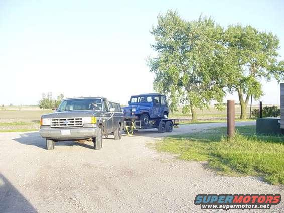Gray Ford Bronco parked on gravel, towing a blue Jeep Wrangler on a trailer