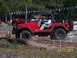 1989 Ford Bronco - Red Jeep Wrangler navigates an off-road obstacle course, with a pickup truck in the background.