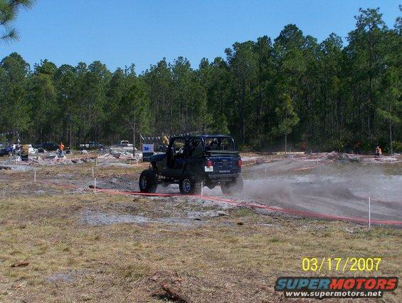 Dark blue off-road vehicle kicking up dust on a dirt track, with trees and other vehicles in the background