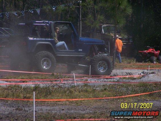 Blue Jeep Wrangler driving through mud, with a red ATV and another vehicle in the background