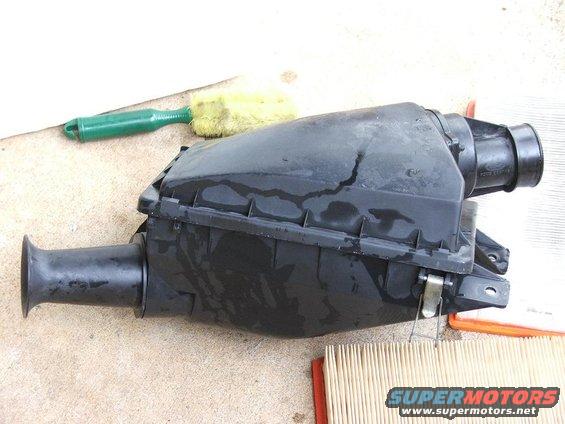 Black plastic air cleaner housing with a green brush and yellow sponge nearby. A new air filter is visible