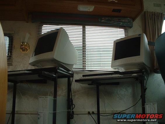 Two CRT computer monitors sit on desks in front of a window with blinds. Computer towers are visible below the desks