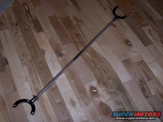 Shiny metal strut tower brace with black mounting brackets on wood floor. Text on brace reads 'Racing'