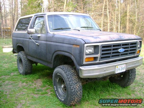 Dark gray Ford Bronco with a white top, lifted suspension, and off-road tires. Front grille and headlights are visible