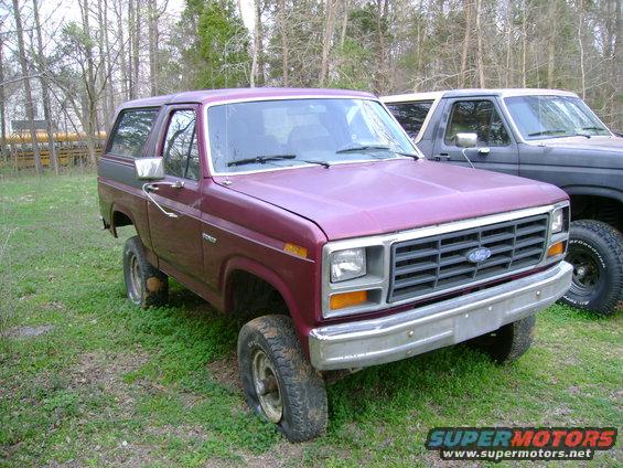 Maroon 1989 Ford Bronco parked in grass with flat tires, another dark SUV behind it