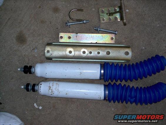 Two white shock absorbers with blue bellows and assorted metal mounting hardware on a concrete surface