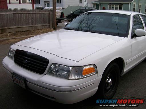 White 1998 Ford Crown Victoria front quarter view. Black grille, clear headlights, orange turn signals. License plate visible