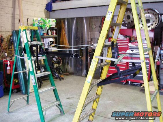 garage with two ladders, one green and one yellow, holding up black car parts. Tool chests and tires are visible in the background
