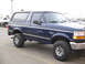 1995 Ford Bronco - This blue 1995 Ford Bronco is shown from a 3/4 front angle, with another white SUV visible in the background.