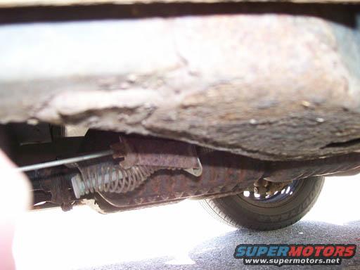 Underside of a car showing rusty frame, exhaust pipe, and a tire. Supermotors.net logo visible