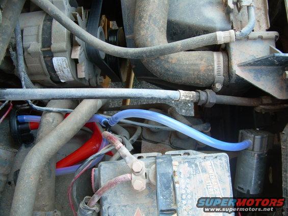 Engine bay showing alternator, battery, and various hoses and wires. Some blue and red tubing is visible