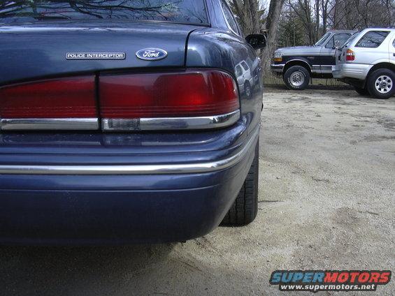 Rear of a blue Ford Crown Victoria Police Interceptor, with a Ford badge and 'Police Interceptor' text. Two other vehicles are parked behind it