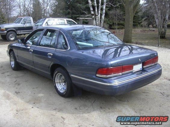 Blue sedan with chrome wheels parked on dirt, rear quarter view. Another truck and SUV are visible