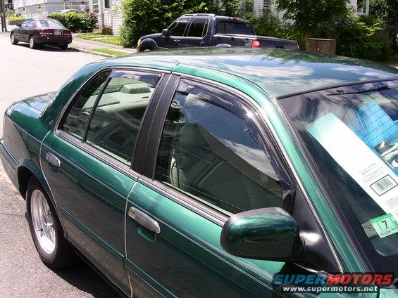 2001 Ford Crown Victoria side view, green paint, chrome trim, window visors. Another car and truck visible in background