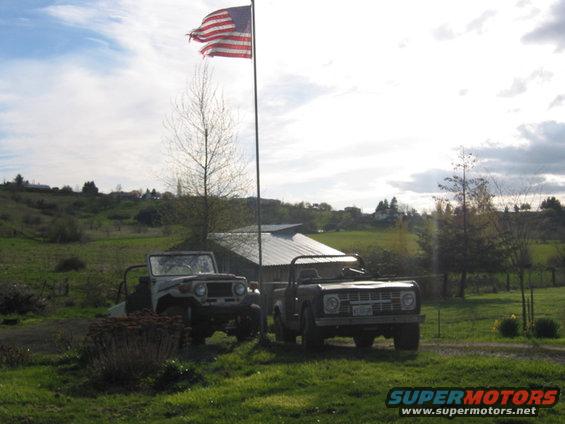 Jeep CJ-5 and a 1973 Ford Bronco parked outside a barn with an American flag flying