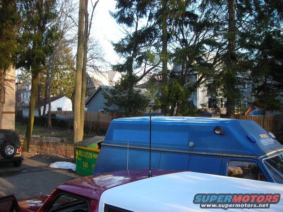 Blue van with a raised roof and antenna, parked next to a dumpster and a red truck