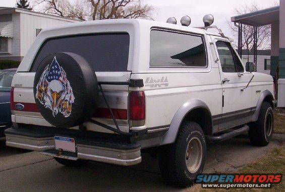 White Ford Bronco, rear quarter view, spare tire cover with eagle and flag graphic, aftermarket wheels, roof lights