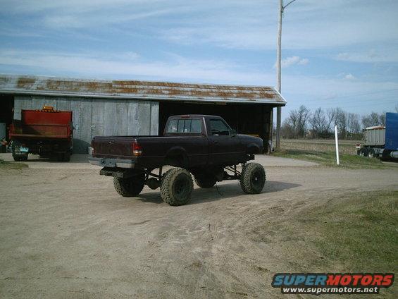 Maroon lifted pickup truck with large tires parked on dirt in front of a barn