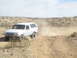 1996 Ford Bronco - A white Ford Bronco navigates a dusty desert trail, kicking up a cloud of dirt behind it.