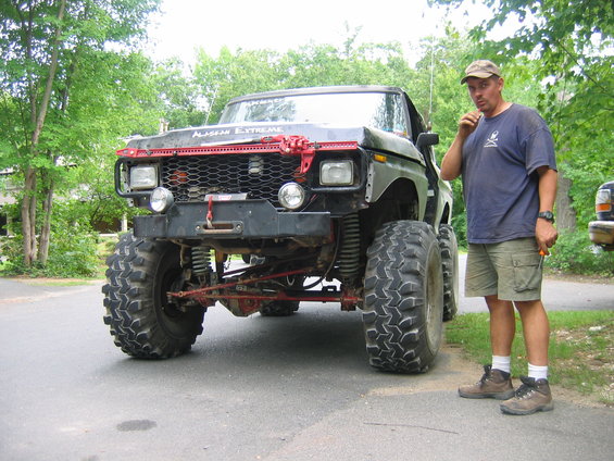 guy standing next to a heavily modified black pickup truck with huge tires and a red grille guard
