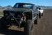 1986 Ford Bronco - This heavily modified pickup truck features oversized tires and a front bumper, with another vehicle following behind in a desert landscape.