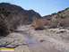 1986 Ford Bronco - A dry creek bed flows through a rocky canyon, with sparse desert vegetation and hills under a clear blue sky.