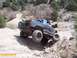 1986 Ford Bronco - Two modified trucks navigate a rocky, off-road trail in a dry, hilly landscape.
