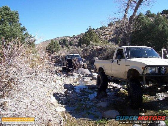 Cream-colored pickup truck with large tires, angled from the front right, driving off-road. Another dark pickup truck is visible behind it