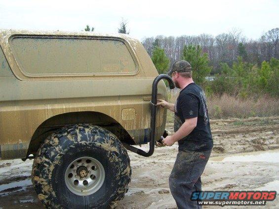 Mud-covered tan Ford Bronco with large off-road tires, rear quarter panel, and a man attaching a roll bar