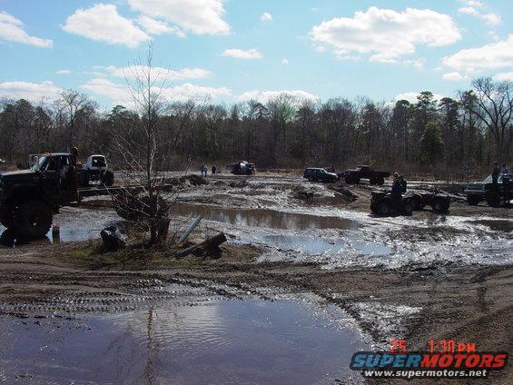Muddy off-road scene with trucks and ATVs. Watermark reads SUPERMOTORS.NET