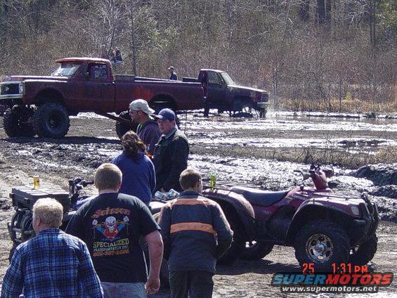 Muddy field with two pickup trucks and a red ATV, people standing around