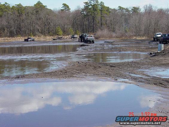 Several trucks are stuck in a muddy area with trees in the background. A water puddle reflects the sky