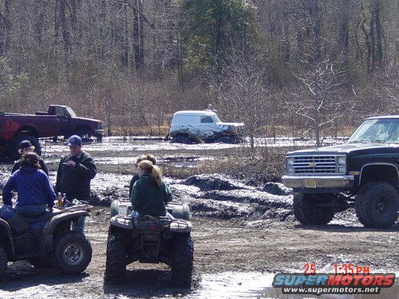 Muddy scene with a lifted black pickup truck in the foreground, people on ATVs, and a maroon truck and white van stuck in mud in the background