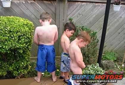 Three young boys urinating outdoors next to a wooden fence and bushes. Watermark reads SUPERMOTORS.NET
