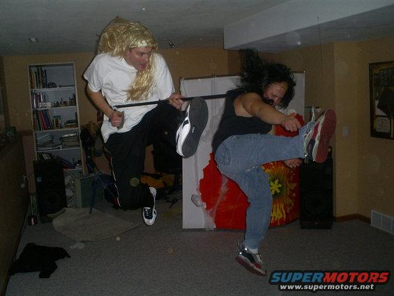 Two people doing karate kicks indoors, one wearing a blonde wig