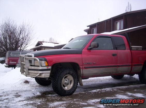 Red Dodge Ram 1500 V8 pickup truck with brush guard in snow. Another red truck with camper shell is behind it
