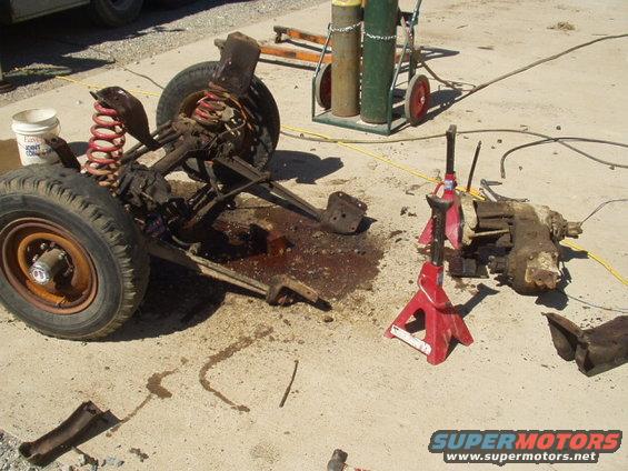 Disassembled front axle and transmission on concrete with red jack stands and welding tanks