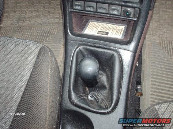 Car interior showing a manual gear shifter, textured floor mats, and a document in a plastic sleeve