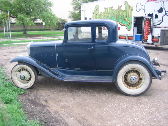 Dark blue coupe with whitewall tires and wire wheels parked on gravel, with a trailer in the background