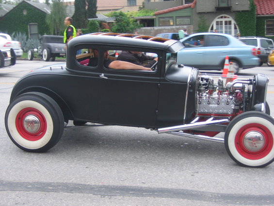 Black hot rod coupe with exposed engine and whitewall tires, driving on a street