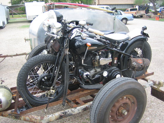 Black vintage Harley-Davidson motorcycle with white seat on a rusty trailer, surfboard on top of a silver trailer behind it