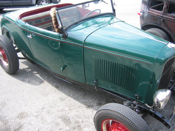 Green hot rod roadster with red wheels and a fur trim on the windshield. Another old car is parked behind it