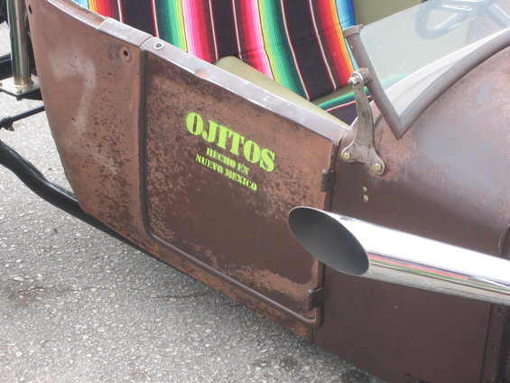 Rusty brown custom hot rod side panel with green text 'OJITOS HECHO EN NUEVO MEXICO' and a chrome exhaust pipe