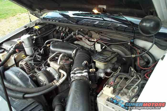 Engine bay with hoses, wires, and engine components visible. Some text on a black air intake tube